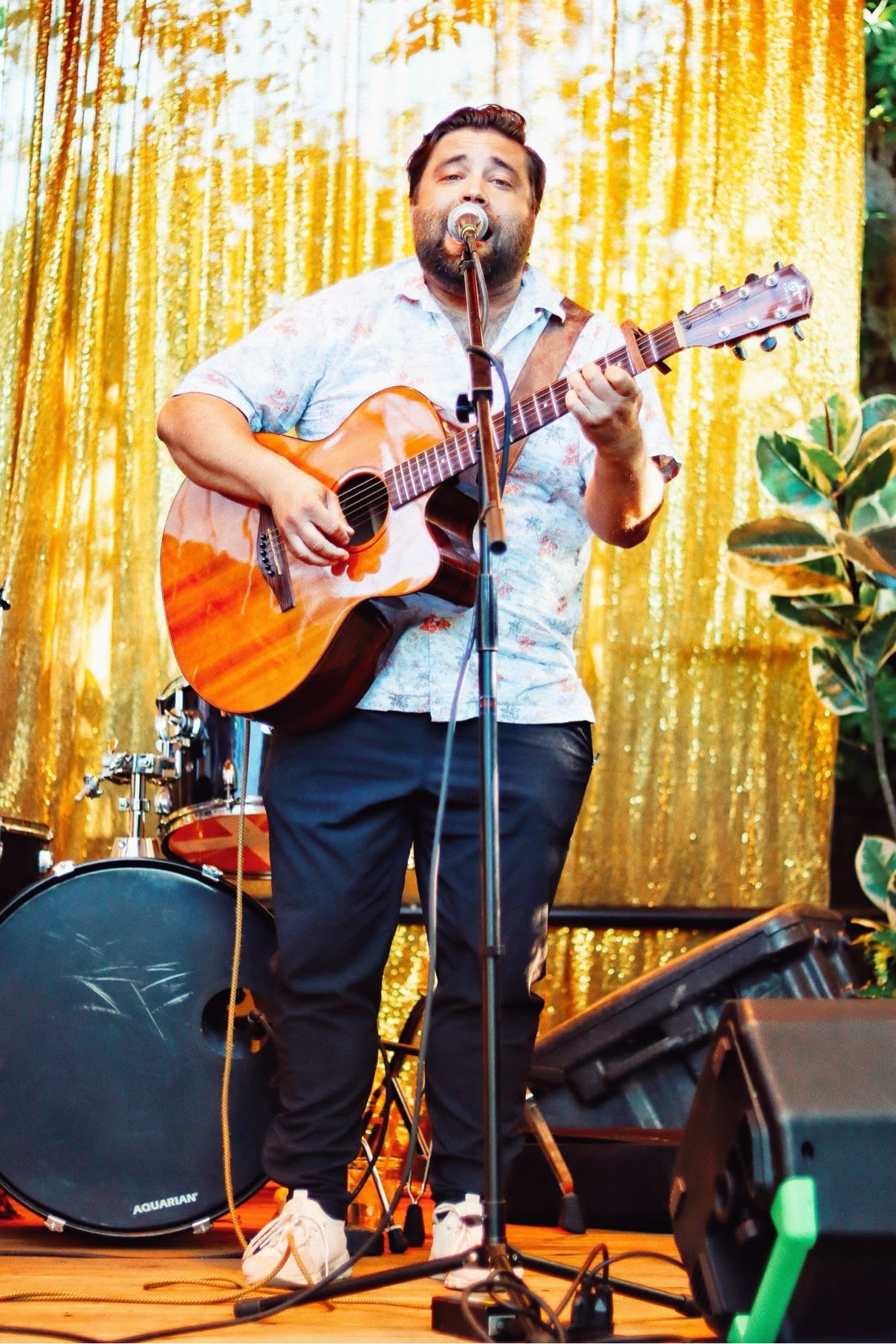 Matt Long performing outdoors in front of a gold sequin curtain, singing into the microphone and playing acoustic guitar with drums behind him