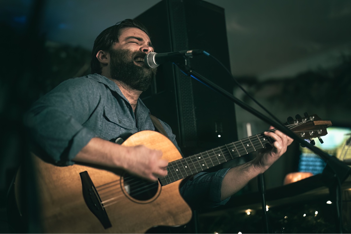 Close-up of Matt Long in a blue shirt singing into a microphone with his eyes closed, playing acoustic guitar on an evening stage