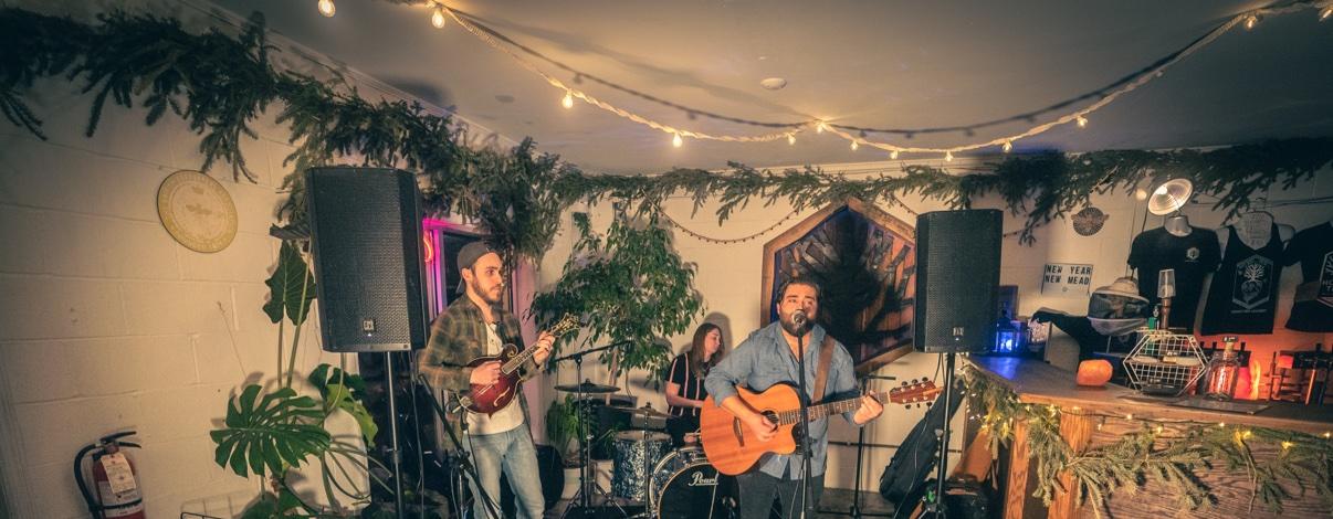 Matt Long singing and playing acoustic guitar with a small band — a mandolin player and a drummer — in a cozy room decorated with greenery garlands and string lights