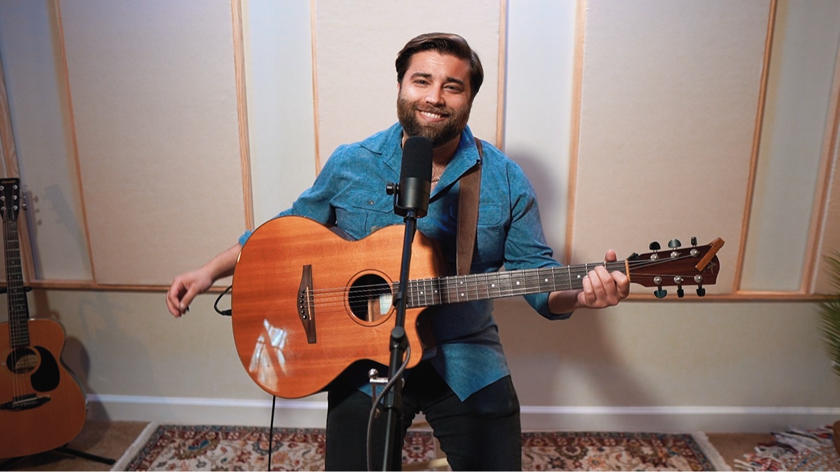 Matt Long smiling and holding his acoustic guitar at a microphone in a warm, paneled house concert room, with a patterned rug and a second guitar on a stand nearby