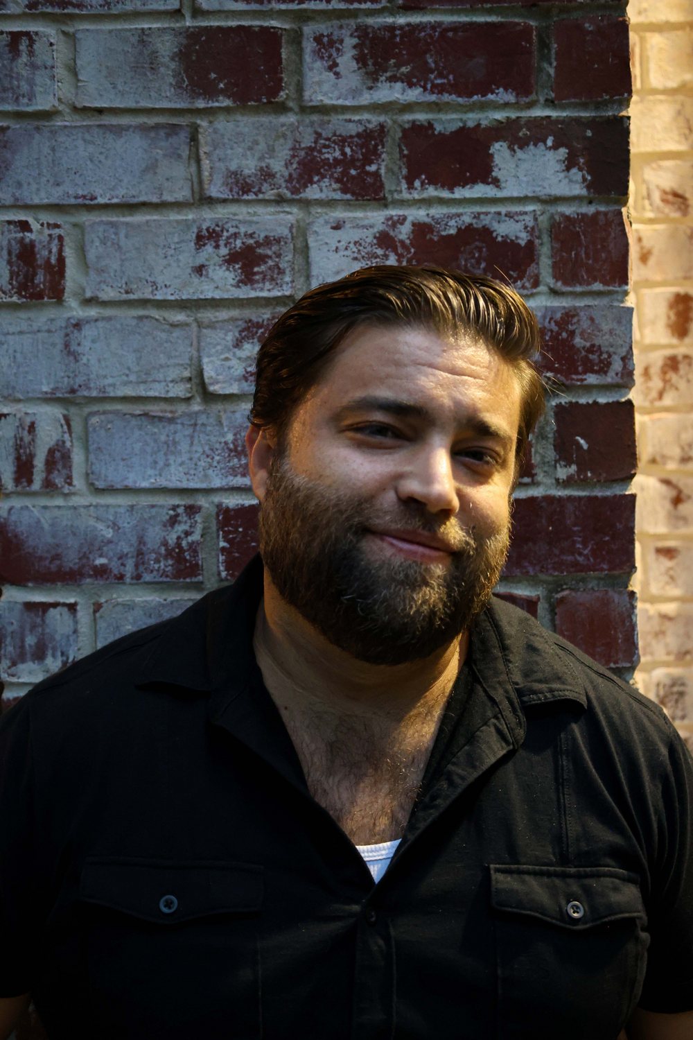 Matt Long smiling in a black shirt against a weathered brick wall