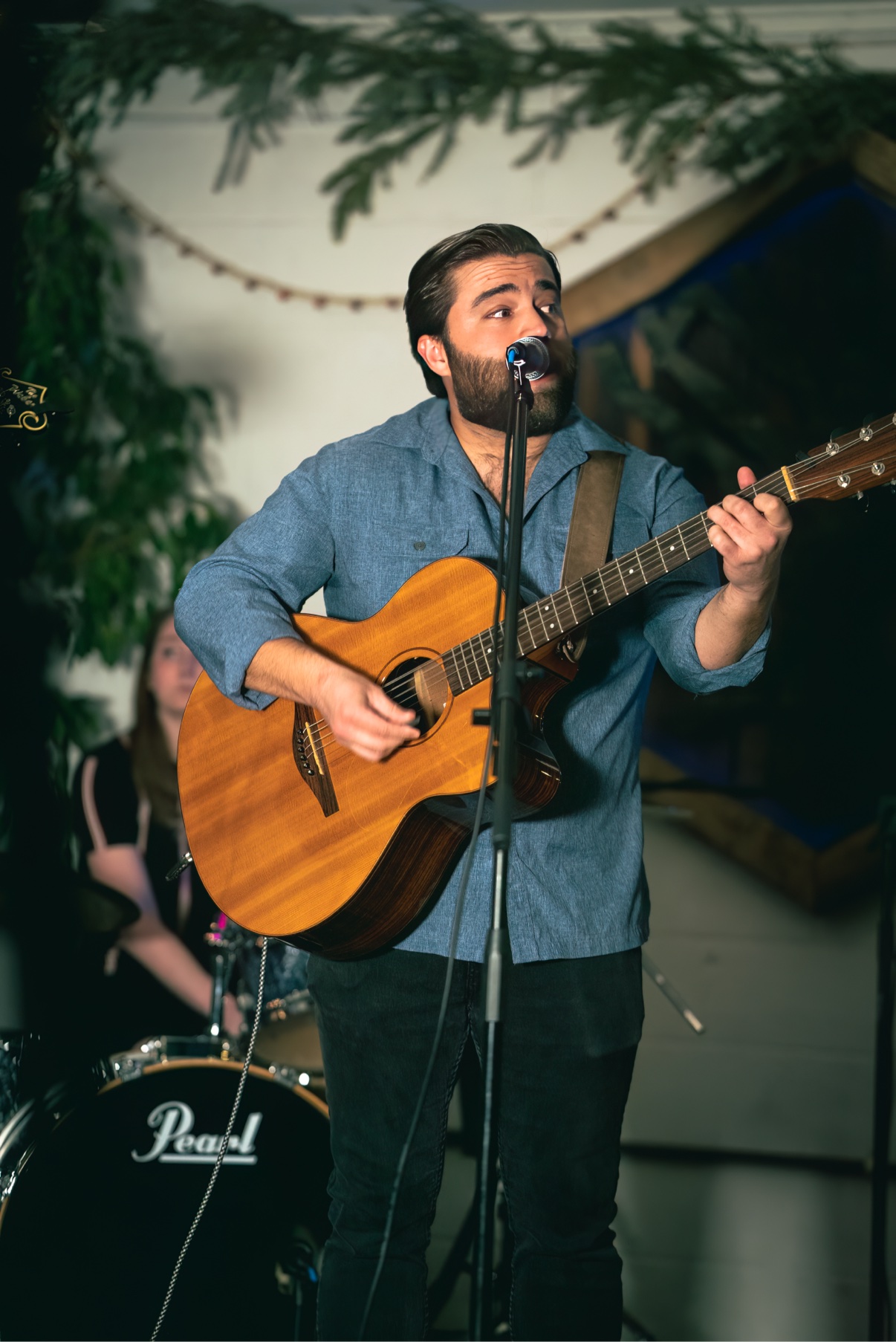 Matt Long singing and playing acoustic guitar at a wedding-style celebration, with greenery garland and string lights overhead and a drum kit behind him