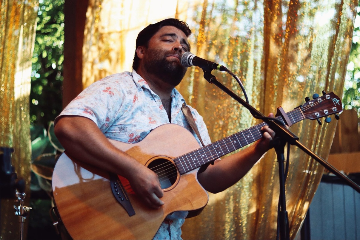 Matt Long singing into a microphone with his eyes closed and playing acoustic guitar outdoors, a gold sequin curtain behind him